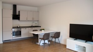 A modern kitchen with white cabinets, a black range hood, and a built-in oven. There is a dining table with two chairs and a flat-screen TV on a white stand. - Top Appartment in Hilden to rent, Lift, Fully furnished with equipped kitchen, (Hilden, Nordrhein-Westfalen, Deutschland)