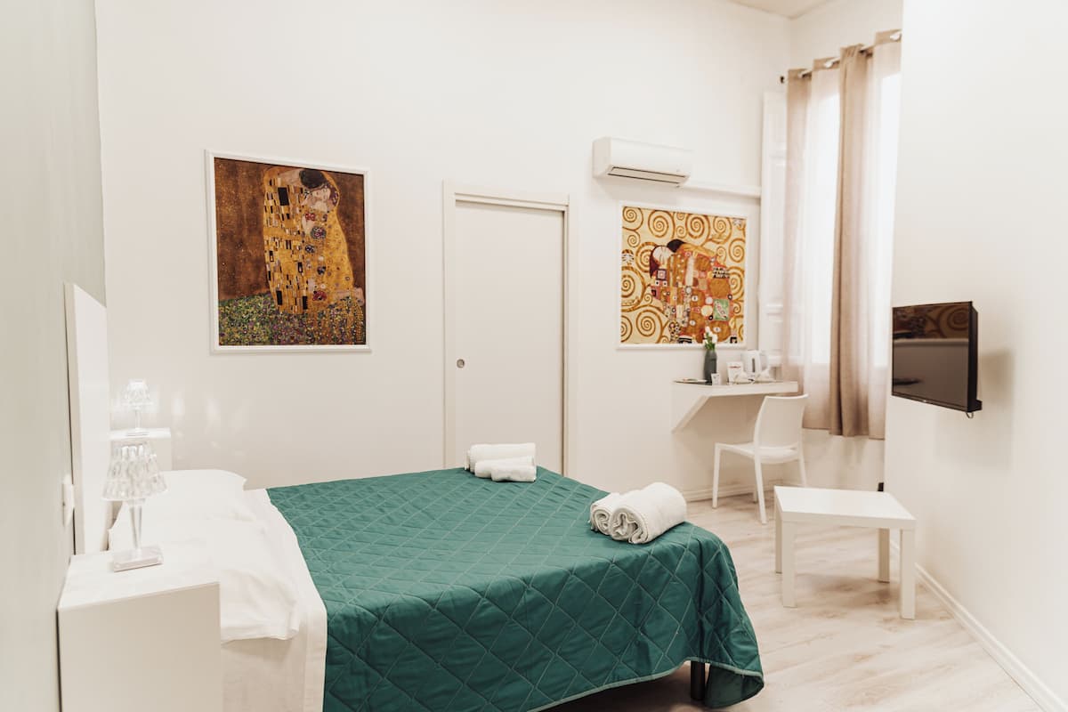 A hotel room with a green bedspread, a painting of a woman, a desk with a chair, and a TV on the wall.