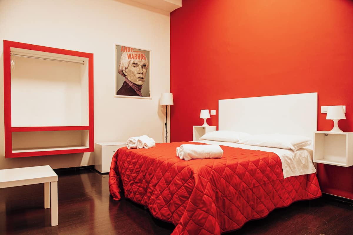 A hotel room with a red wall, a bed with white bedding, a red quilted bedspread, a white bedside table, a white nightstand, and a poster on the wall.