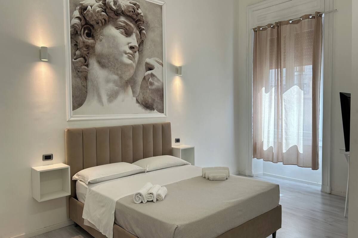A modern bedroom with a large bed, a headboard, bedside tables, a wall-mounted artwork, and a window with curtains.