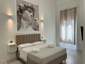 Un dormitorio moderno con una cama grande, cabecero, mesitas de noche, una obra de arte en la pared y una ventana con cortinas.