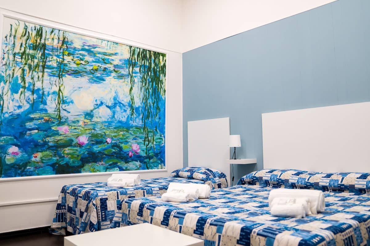 A hotel room with two beds, a large painting of water lilies on the wall, and a bedside table with a lamp.