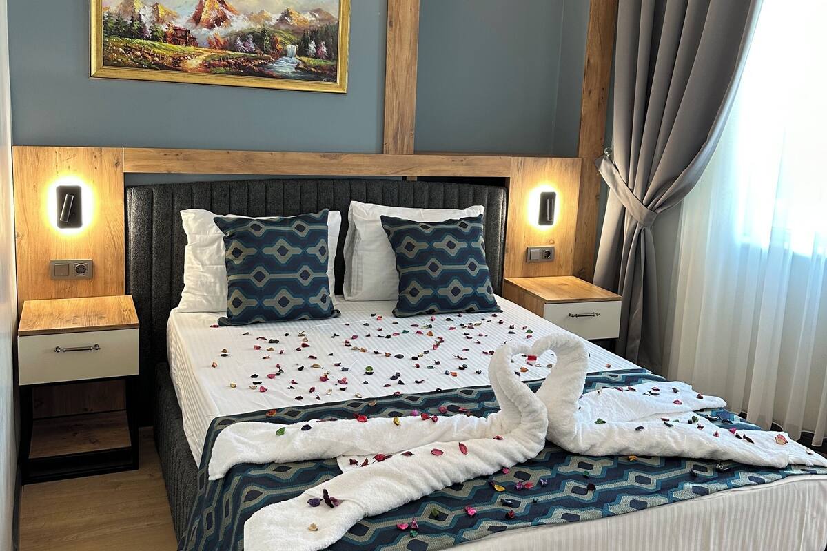 A neatly made bed with patterned bedding, two bedside tables with lamps, a painting on the wall, and a window with curtains.