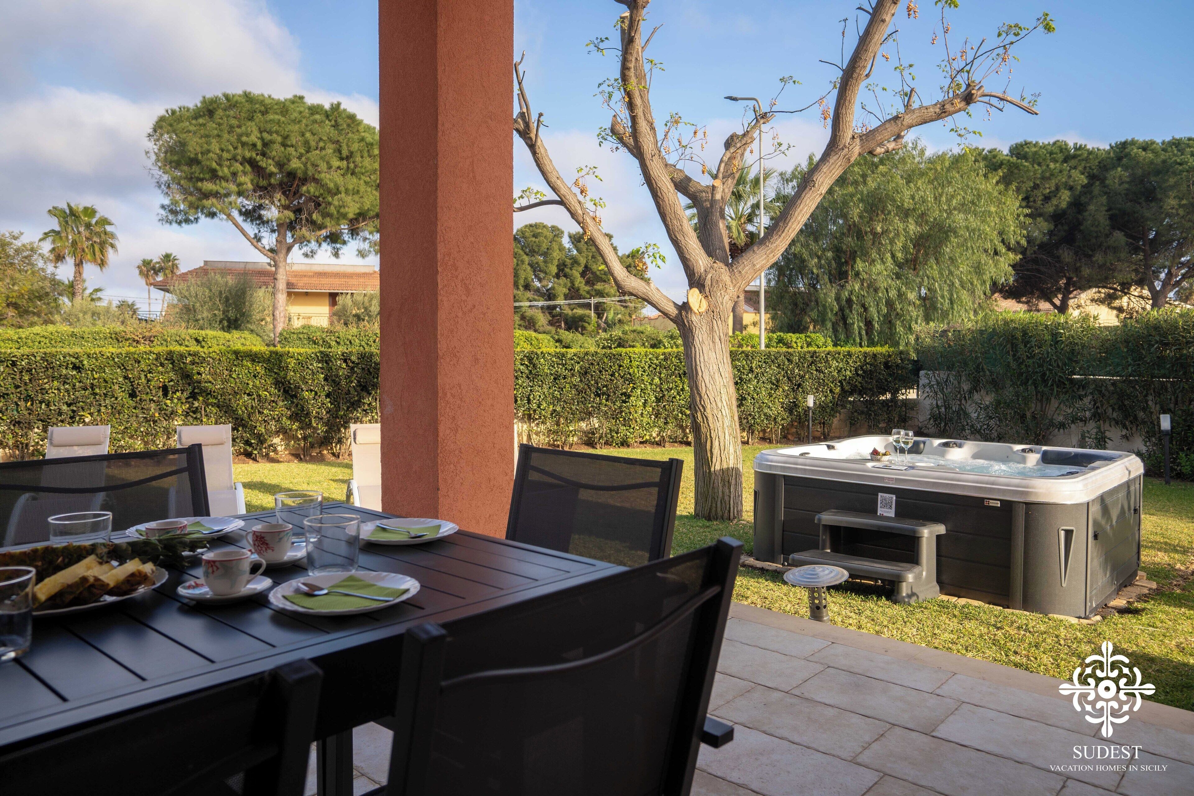 An outdoor dining area with a table set for two, surrounded by chairs, and a hot tub in the background.