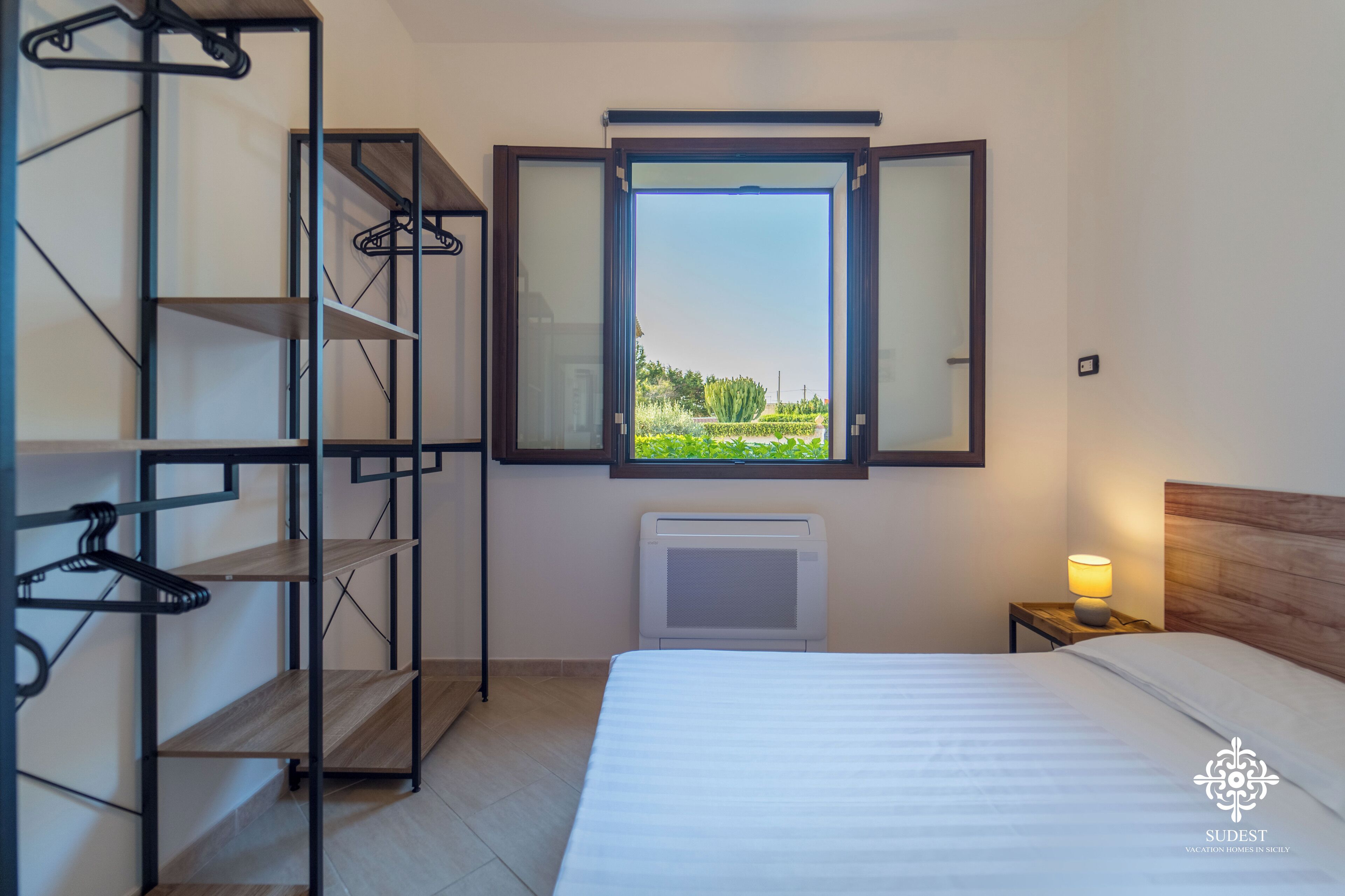 Ett hotellrum med en säng, ett fönster med utsikt över grönområden, ett element och en hylla med klädhängare.