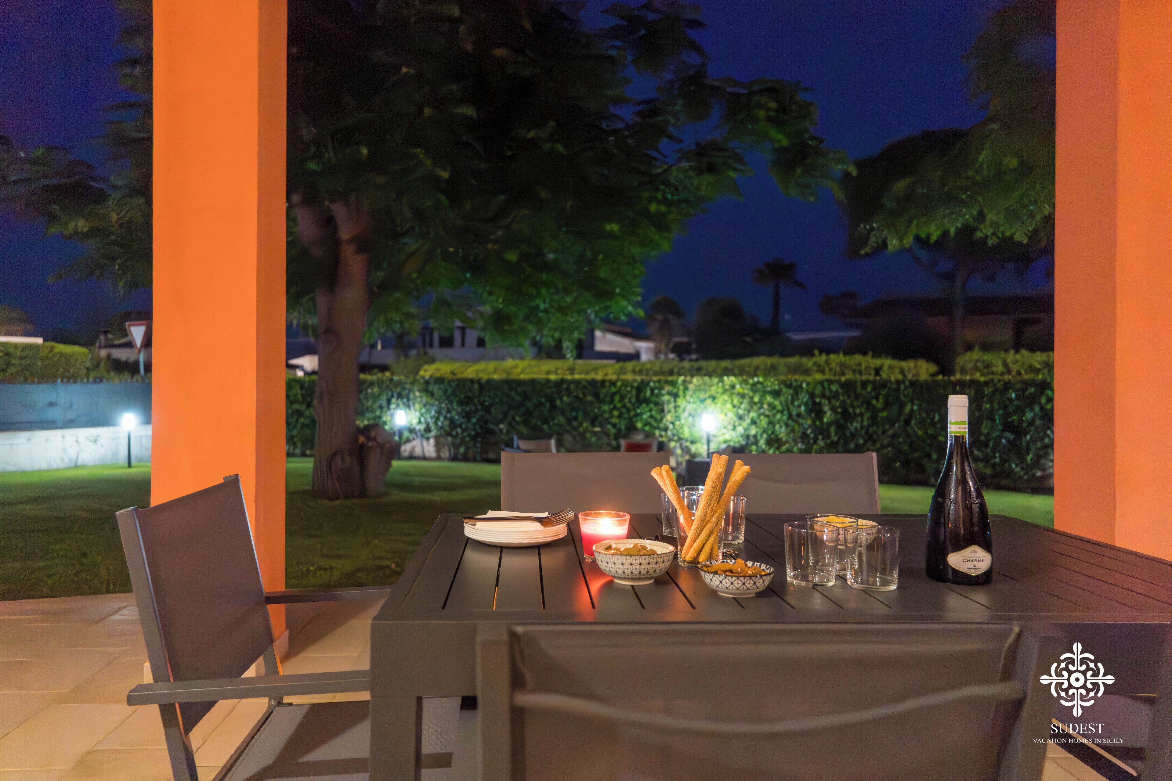 An outdoor dining table set with plates, breadsticks, glasses, and a bottle of wine, surrounded by chairs and a candle, with a hedge and trees in the background.