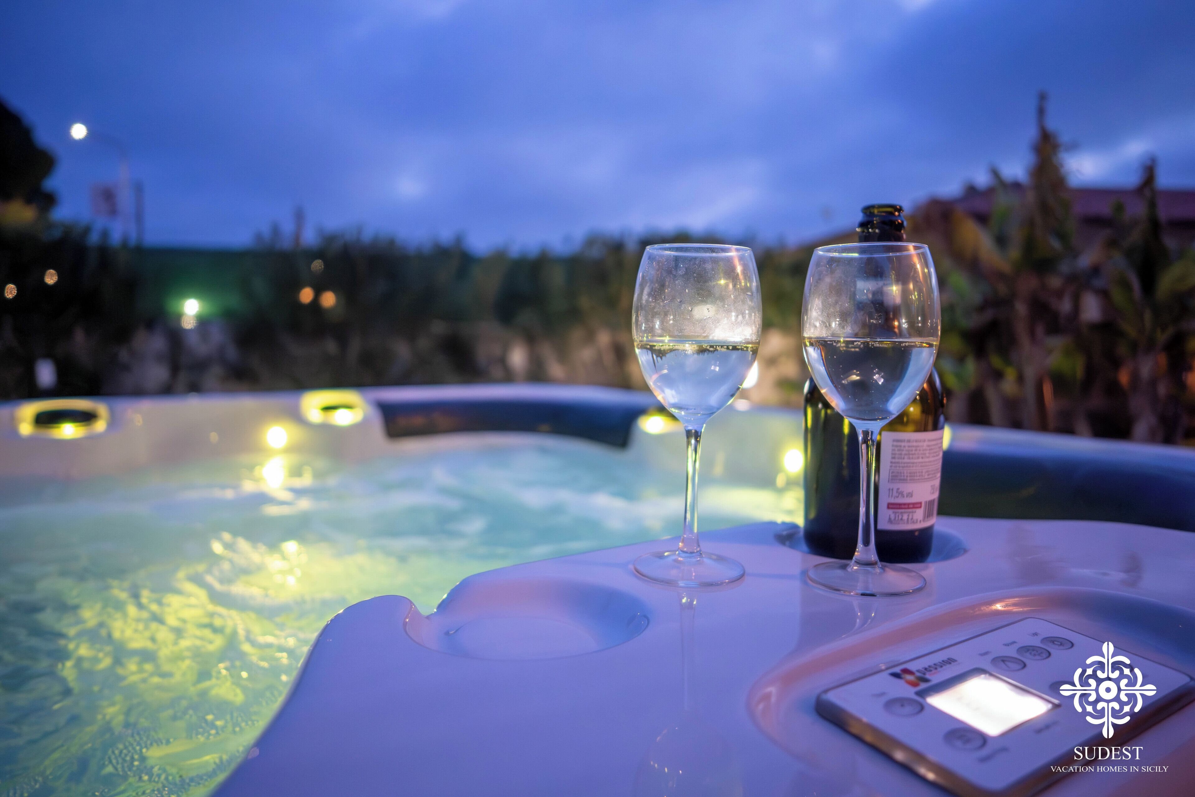 A hot tub with two glasses of white wine and a bottle, surrounded by a pool and trees.