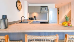 Een moderne keuken met een centraal eiland, een magnetron en een koelkast. Er hangt een klok aan de muur en op het aanrecht staat een fruitschaal.