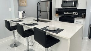 A modern kitchen with white cabinets, a stainless steel refrigerator, a microwave, and a gas stove. There are two black bar stools and a coffee maker on the counter.