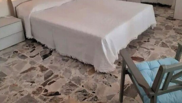 Ein Schlafzimmer mit Steinfußboden, einem Bett mit weißer Bettwäsche, einem Nachttisch und einem Sessel mit blauer Sitzkissen.