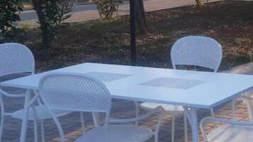 Uma área de estar ao ar livre com uma mesa e cadeiras brancas, cercada por árvores e um poste de luz.