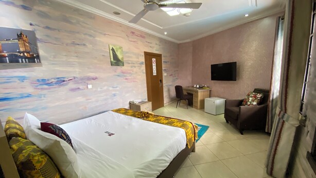 A hotel room with a bed, a desk, a chair, a television, and a ceiling fan. - Bays Villa (Koforidua)