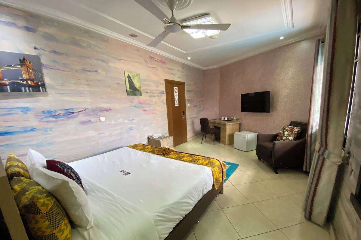Ein Hotelzimmer mit Bett, Schreibtisch, Sessel, Fernseher und Deckenventilator.
