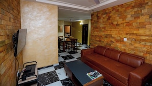 A room with a brown leather sofa, a black coffee table, a TV mounted on the wall, and a ceiling fan. - Bays Villa (Koforidua)
