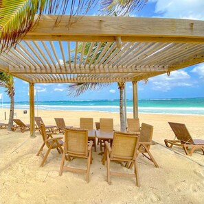 Un cabanon de plage en bois, équipé d’une table et de chaises, avec vue sur la plage et l’océan.