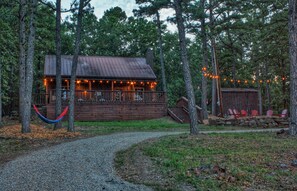 A rustic cabin with a porch, surrounded by trees and a gravel path.