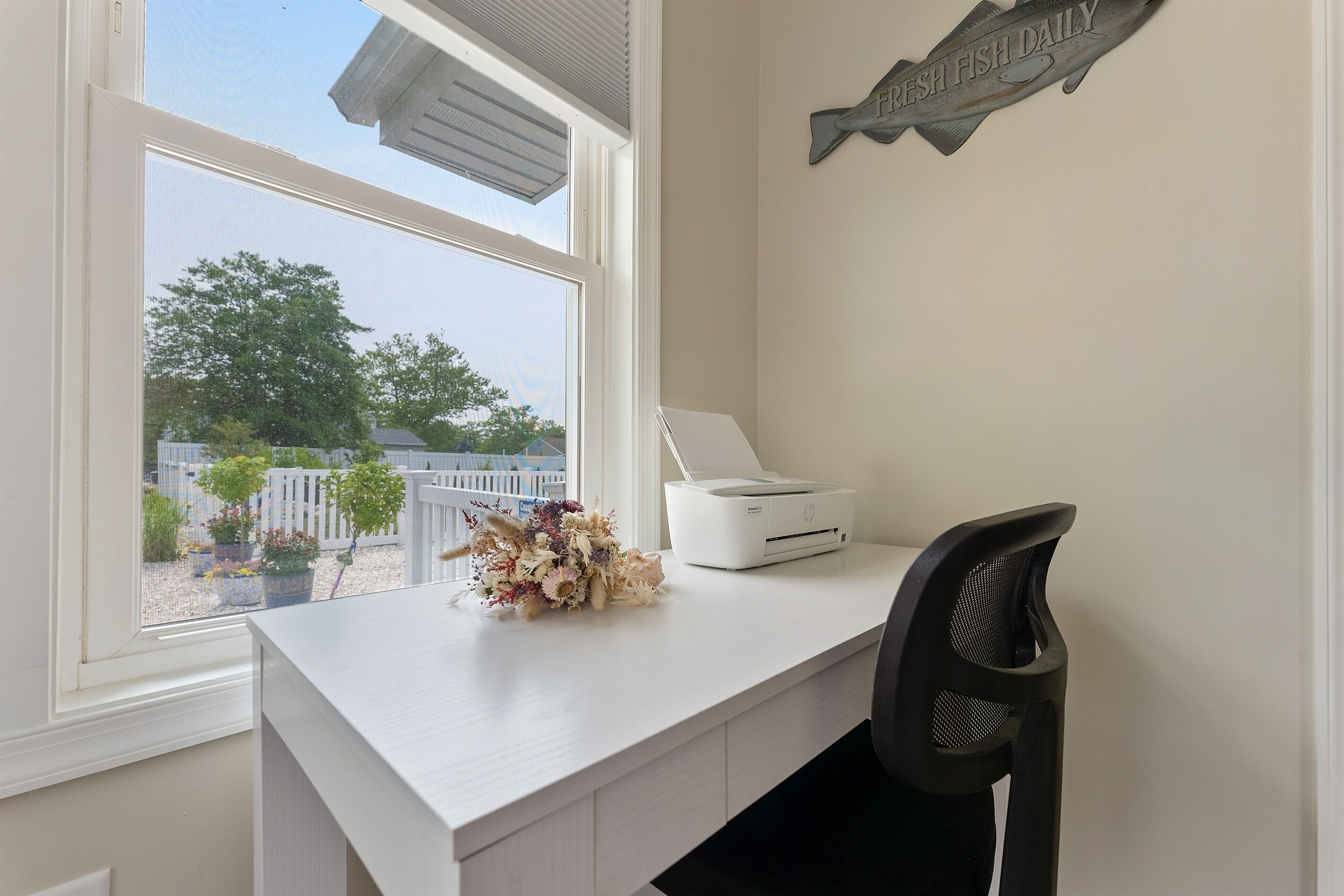 A white desk with a printer, a black chair, and a window with a view of a garden and trees.