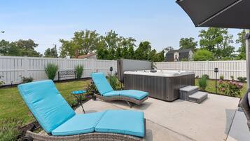 A backyard with two wicker lounge chairs with blue cushions, a hot tub, and a patio umbrella.