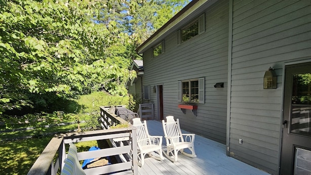 A covered deck with white chairs and a bench, a building with a door and windows, and a tree-lined background. - Bolton Landing - 4 Bdrm Vacation Rental (Bolton Landing)