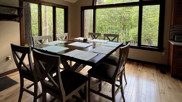 Un espace repas comprenant une grande table, des chaises et une vue sur les arbres, visible à travers une grande fenêtre en arc de cercle.