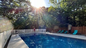 Une piscine avec une terrasse en bois, une clôture blanche et un espace abrité équipé de chaises longues.
