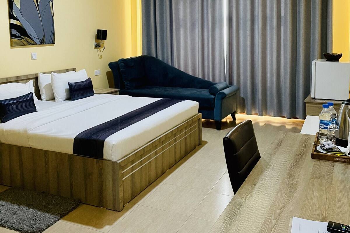 A hotel room with a bed, a desk with a chair, a sofa, a curtain, and an air conditioning unit.