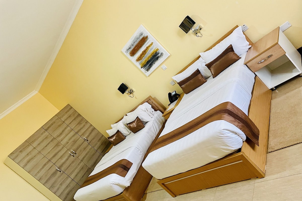 A hotel room with two beds, a wardrobe, a bedside table, a lamp, and a painting on the wall.