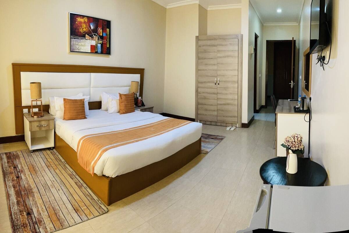 A hotel room with a large bed, bedside tables, a desk, a chair, a wardrobe, and a painting on the wall.