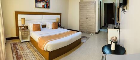 A hotel room with a large bed, bedside tables, a desk, a chair, a wardrobe, and a painting on the wall.