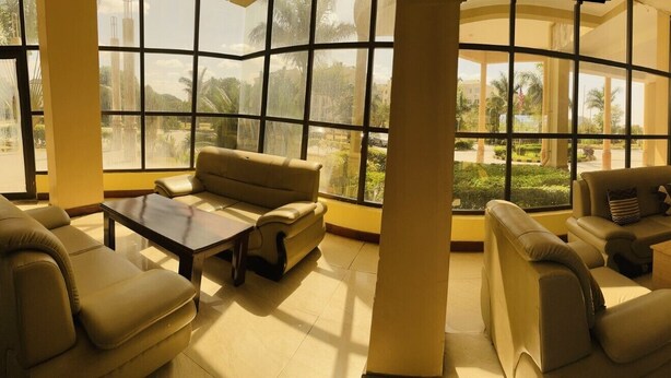 A spacious lounge with large windows, a sofa, a coffee table, and a small table with a lamp. - NASHERA HOTELS DODOMA (Dodoma)