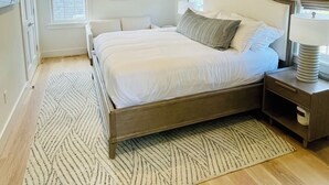 A bedroom with a sloped ceiling, a bed with white bedding, a bedside table with a lamp, and a patterned rug. - Charming new construction 4br cottage in town (Nantucket)