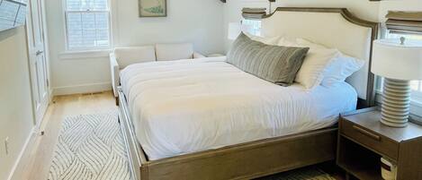 Un dormitorio con techo inclinado, una cama con ropa de cama blanca, una mesita de noche con lámpara y una alfombra estampada.