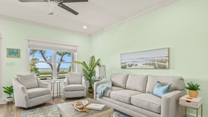 A living room with a light green wall, a beige sofa, two armchairs, a coffee table, and a view of the outdoors through a large window. - Scenic Sandpiper by the Shore (Long Beach)