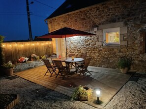 A stone building with a wooden deck, outdoor table and chairs, string lights, and potted plants. - Le Boiselande - Landebia (LANDEBIA)