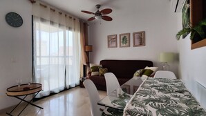 A living room with a dining table, a sofa, a ceiling fan, and a window with sheer curtains.
