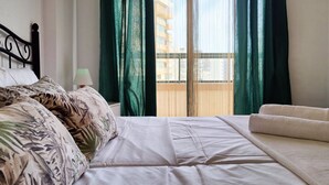 A hotel room with a bed, a sofa, a nightstand, a lamp, and a view of a building through a window with green curtains.