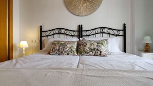 A neatly made bed with a white comforter and two floral pillows, a round wall decoration above the bed, and two bedside lamps on either side.