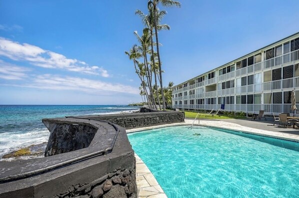 A pool with a view of the ocean, a building with balconies, and palm trees.