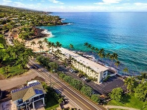Vista aérea de una zona costera con un edificio junto a la playa, que incluye un estacionamiento y paneles solares en una estructura cercana.
