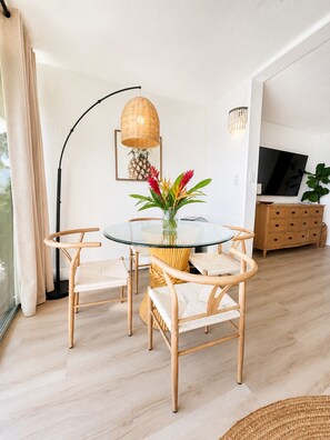 A modern dining area with a glass table, wicker chairs, a woven pendant light, and a wooden cabinet with a flat-screen TV.