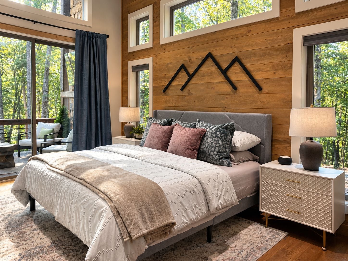 A bedroom with a large bed, a nightstand, a dresser, and a view of trees through the windows.