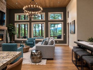 A spacious living room with a fireplace, large windows, a chandelier, and a dining area. - 4 Kings| No Bunks| Serene Views| Sauna| EV| Pets (Blue Ridge)