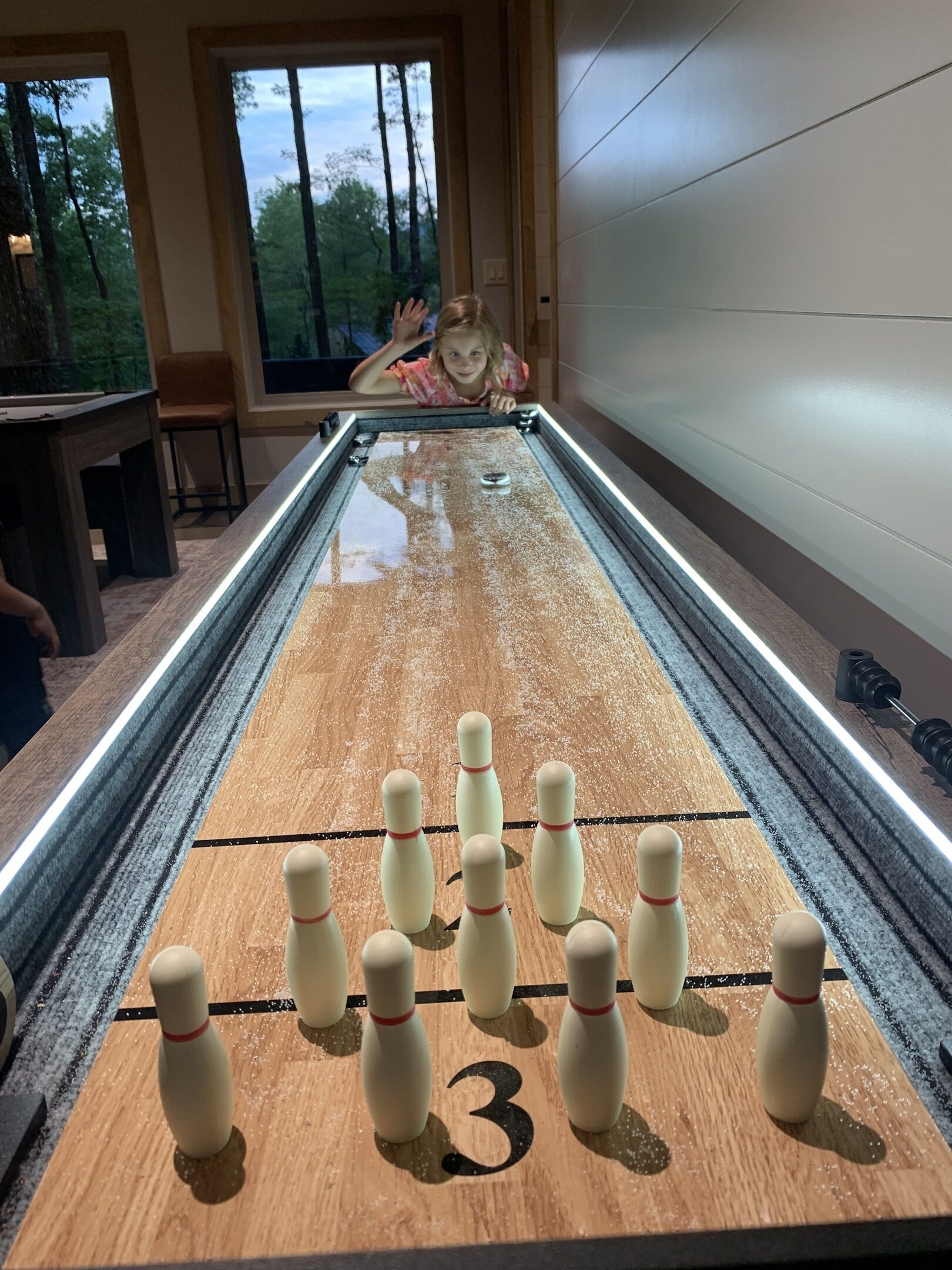 A wooden shuffleboard table with bowling pins set up for a game, a person in the background, and a view of trees through a window.