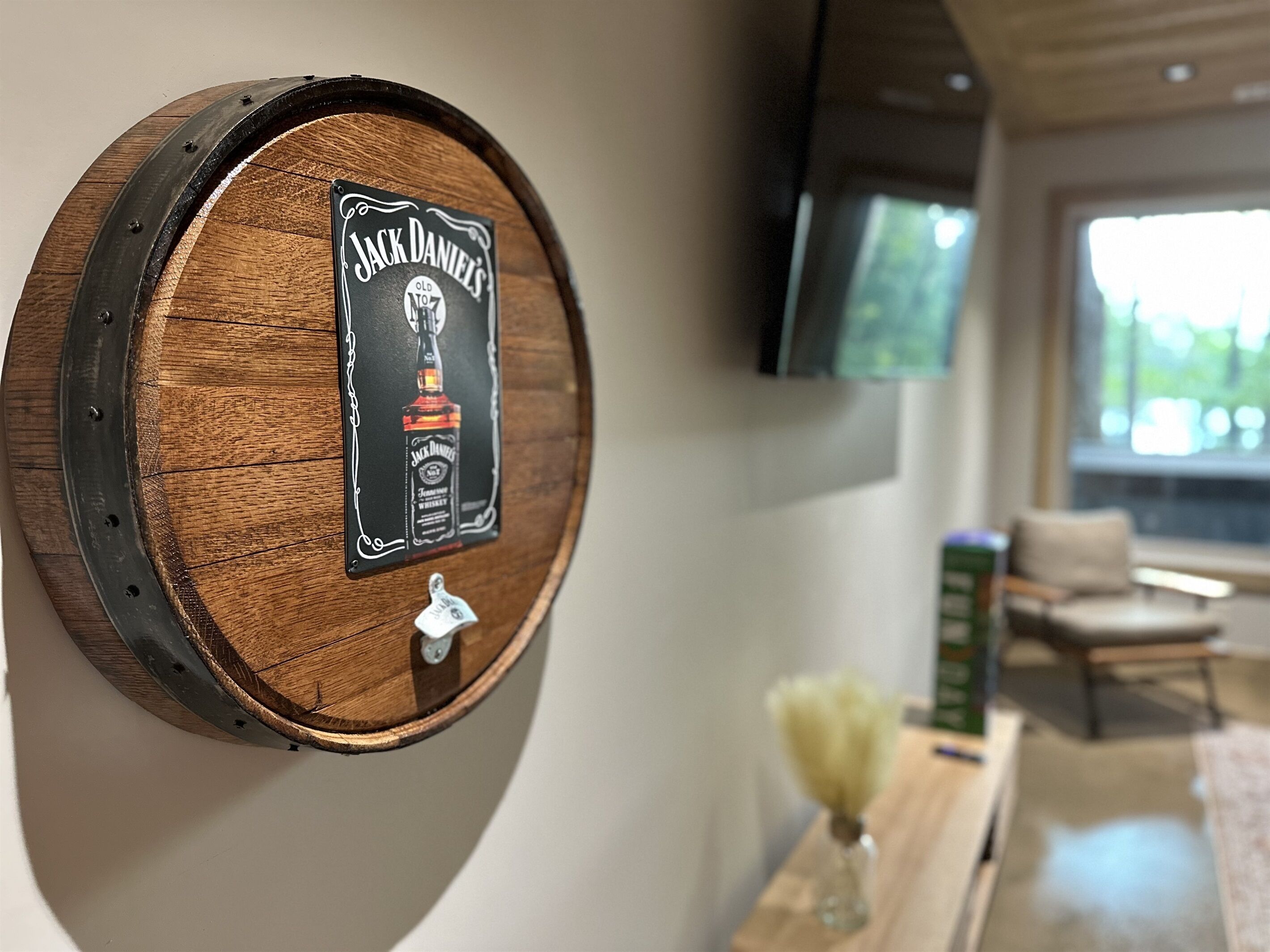 Eine Wanddekoration aus Holzfässern mit Jack Daniel's Etikett, ein angebrachter Fernseher, eine Vase mit Blumen und ein Sessel in einem modernen Wohnzimmer.