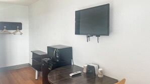 A small room with a flat-screen TV mounted on the wall, a wooden table with a remote control, a small speaker, and a trash bin.
