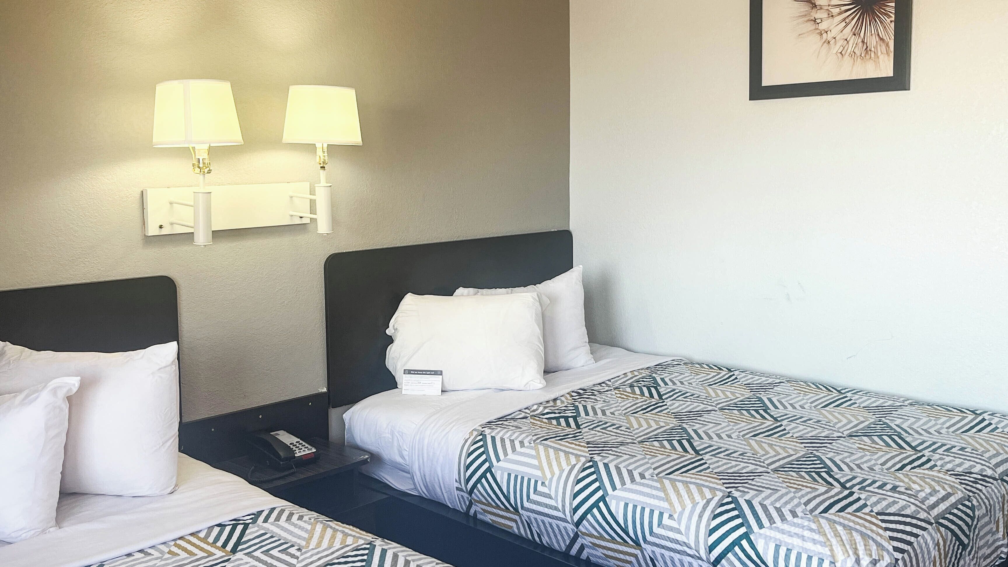 A hotel room with two beds, each with white bedding and a patterned blanket. There are two wall-mounted lamps with white shades, a framed picture on the wall, and a telephone on the nightstand.