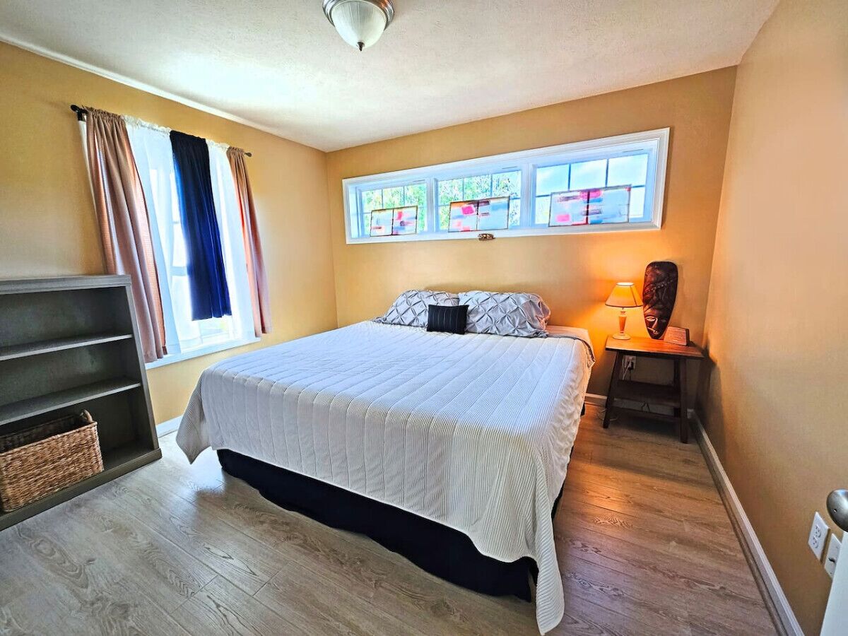 A bedroom with a bed, a nightstand, a lamp, a window with curtains, and a shelf with baskets.