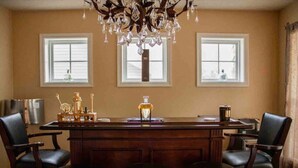 A bar area with a wooden bar, two black leather chairs, a chandelier, and two windows with blinds.
