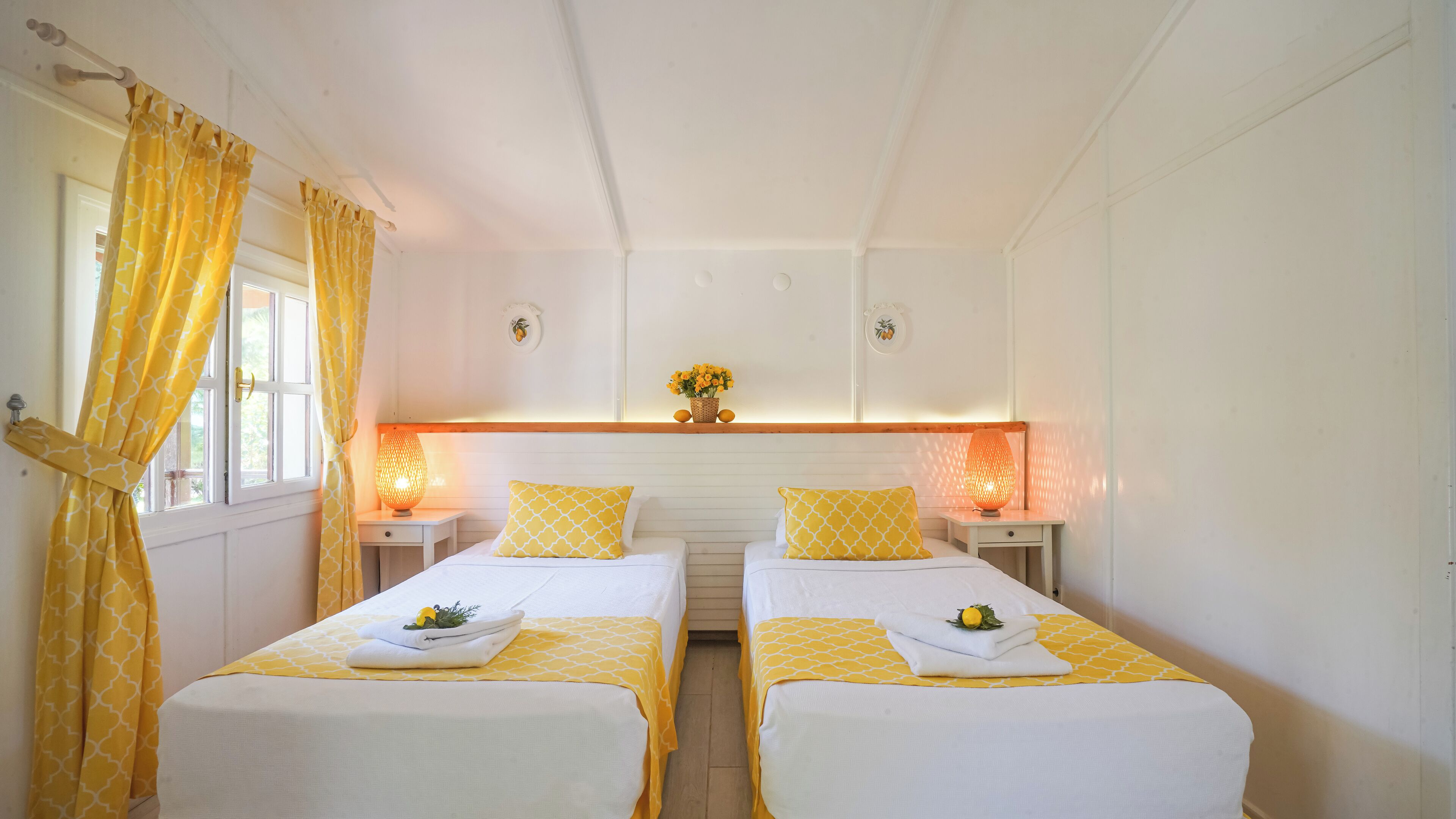 Una habitación con dos camas, cada una con colcha de diseño amarillo y blanco, una mesita de noche con lámpara y una ventana con cortinas amarillas.