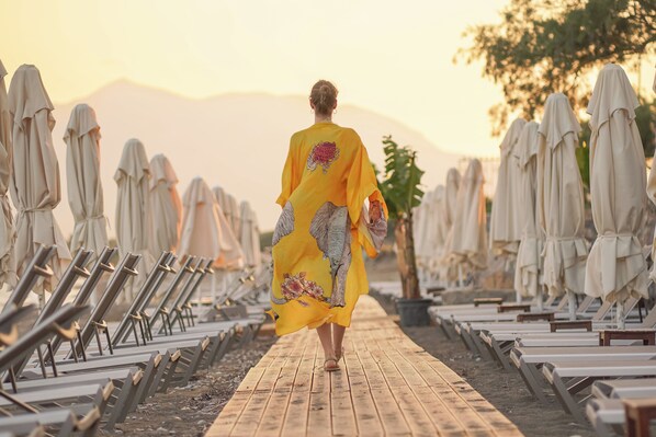 A person walking along a wooden pathway lined with beach chairs and umbrellas, with mountains in the background. - Dirik Surf & Beach Club Hotel Datça (Datça)